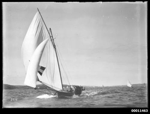 18-foot skiff with dark pennant and lighter cross as sail insignia runs down wind on Sydney Harbour with Manly in far distance