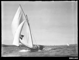 18-foot skiff with dark pennant and lighter cross as sail insignia runs down wind on Sydney Harbour with Manly in far distance