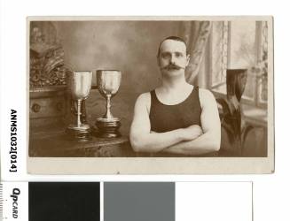 A man in a swimming costume with elaborate moustache, with arms crossed, and standing next to two trophies