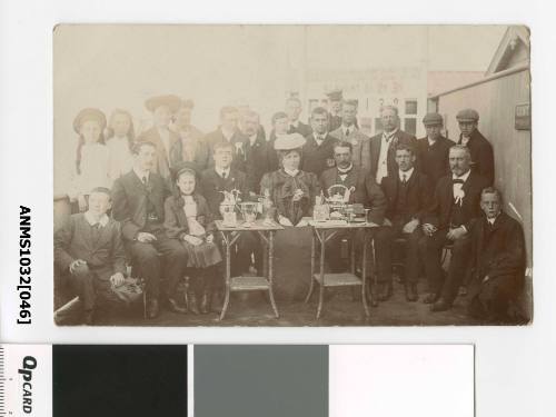 A group of men and women around two table, on which are numerous trophies