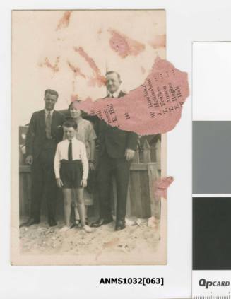 Postcard featuring a black and white photograph of two men, a woman and a young boy, standing on sand in front of a wooden fence