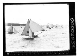 AUSTRALIA (Union jack) leads the 18-footer fleet including MASCOTTE (Diagonal Cross), ROSETTA (Red and blue ball) and MISSISSIPPI (Maltese Cross).