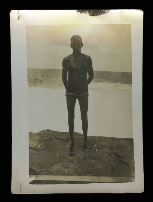 Man wearing a dark coloured swimsuit, who is standing on a large rock, with the waves crashing in the ocean behind him