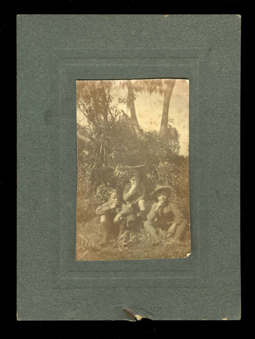 Black and white photograph of three young boys sitting in front of a tree and leafy tree branches, possibly boy scouts