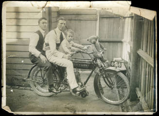 Two men and a young boy (Tom Wild) on a motorbike, with a teddy bear sitting on the handle bars