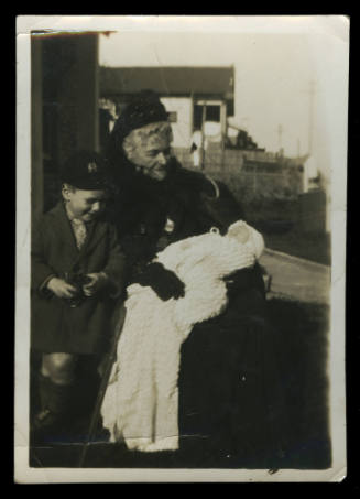 An elderly woman in a black outfit, with a young boy beside her, and holding a baby wrapped in a white blanket