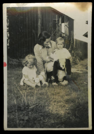 Woman kneeling on the grass next to two young children, with a toy dog also sitting on the grass, and a shed is in the background