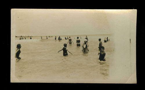 Black and white photograph of numerous people swimming and standing in the water at the beach