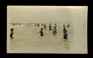 Black and white photograph of numerous people swimming and standing in the water at the beach