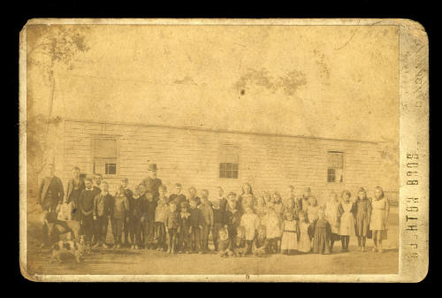 Black and white photo on thick cardboard of a school group, with teachers and two dogs, lined up in front of a long wooden building with three windows