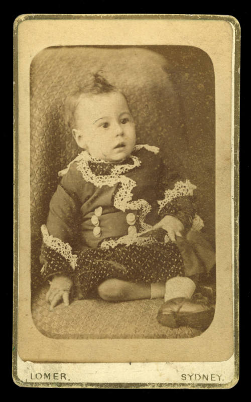 A baby sitting upright, wearing a jacket with sections of white lace sewn onto it, and white socks and shoes