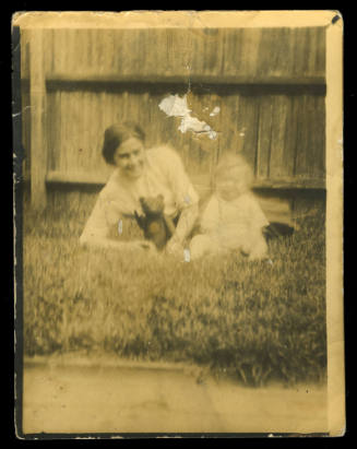 Black and white photograph of Beatrice Kerr laying on the grass in front of a fence, next to a young baby, holding a small teddy bear