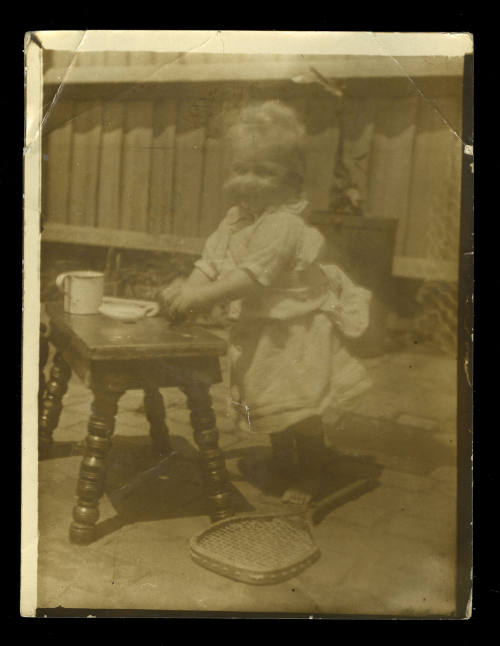 A baby standing at a small table holding a cup and saucer, with a tennis racket on the floor next to her feet