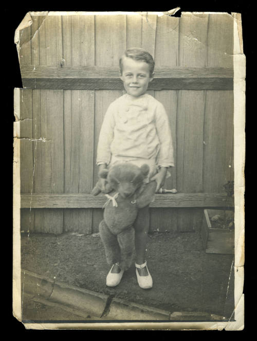 Young boy in a white jacket and shoes, holding a teddy bear in front of him by the arms