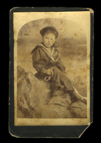 Young boy in a sailor outfit, holding onto a rope, and sitting on a rock