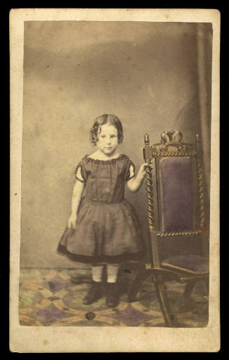 Young girl standing, holding onto a wooden chair, wearing a dress