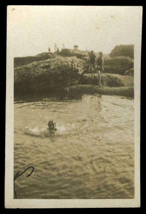 Young boy swimming in the water next to the shoreline, with several people standing on large rocks beside the water