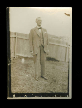 Man standing on lawn in a garden, with a wooden fence in the background, wearing a suit and tie