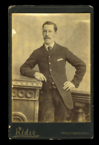 Man with a moustache leaning on a column, with his other hand on his hip