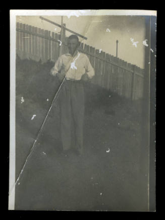 Man standing on the lawn in front of a wooden fence, with his hands on his belt, wearing a pale coloured jumper