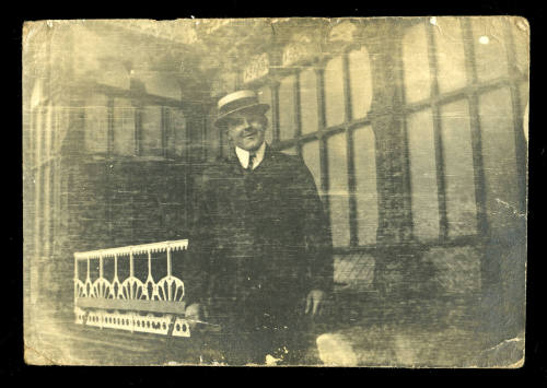 Man wearing a hat and dark coloured suit, standing in front of a white railing and large windows of a building