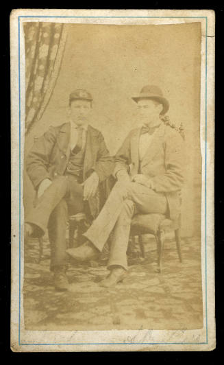 Black and white photograph of two men sitting beside each other in chairs, both with their legs crossed, and wearing hats and suits