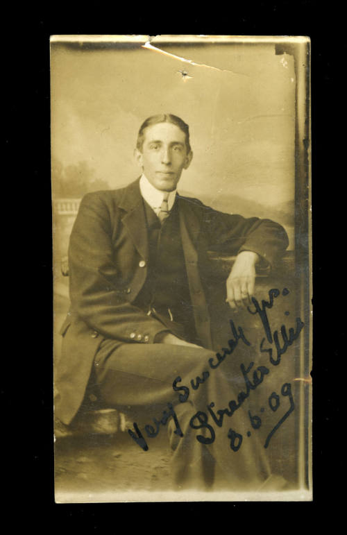 Man sitting in a chair with his arm resting on a column, and wearing a suit