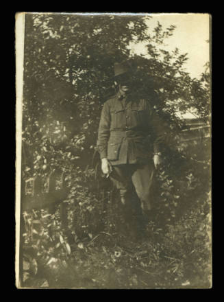 Man posing for the camera in full army uniform, next to a gate, and in front of a bushy tree