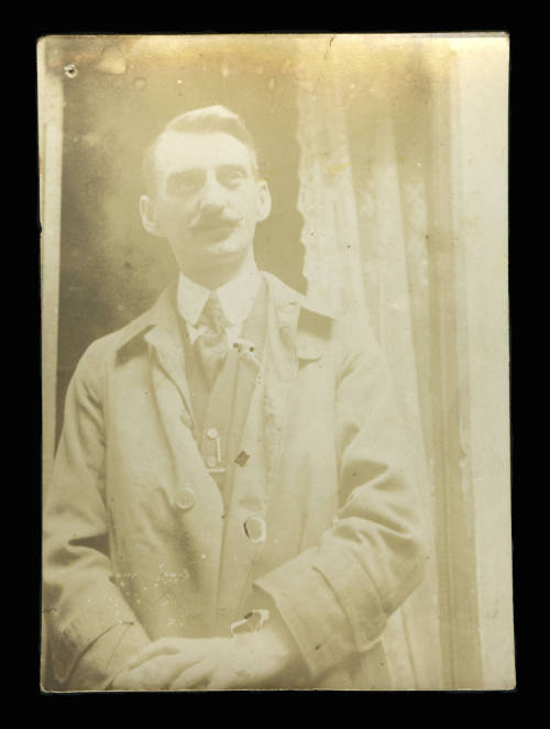 Man standing in front of a doorway with a lace curtain, wearing a jacket, and with his hands together