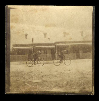 Black and white photograph of two people riding bikes on the road in front of two small houses with picket fences