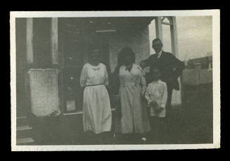 Black and white photograph of four people, two women on the left, a man on the right, and a young boy in the front of the group, standing in front of the verandah of a house
