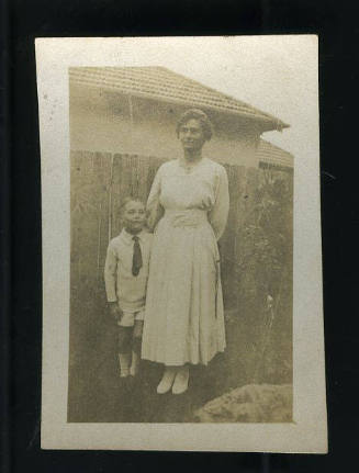 Black and white photograph of Beatrice Kerr standing next to a small boy in front of a wooden fence, with a house in the background