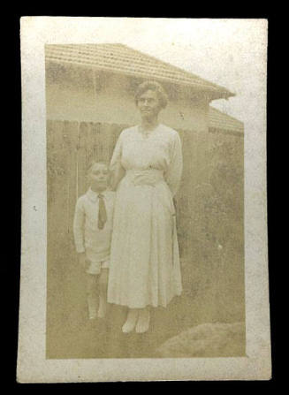 Black and white photograph of Beatrice Kerr standing next to a small boy in front of a wooden fence, with a house in the background