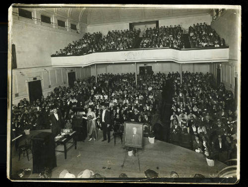 A large gathering of people in a large room, with people sitting at ground level and on a balcony, with people on stage