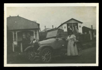 Woman standing in front of a car, with two houses visible in the background