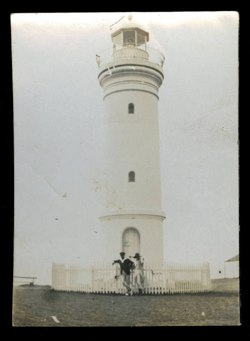 A lighthouse, with a white picket fence around the base of the lighthouse, and two men standing at the front gate