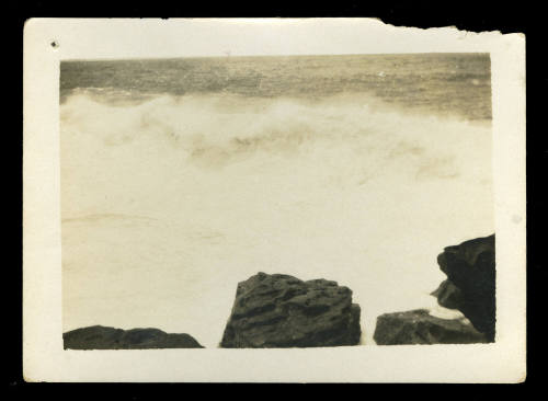 Black and white photograph of the ocean, with rocks in the foreground, and waves crashing in the background