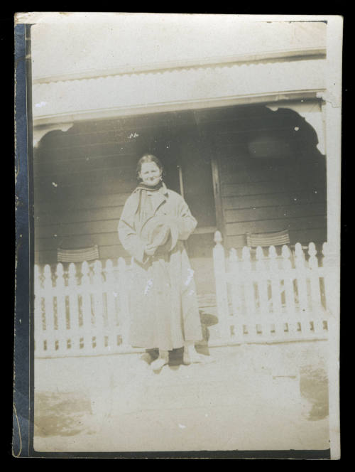 Black and white photograph showing a woman standing in front of a white picket fence and a house entrance, wearing a large jacket