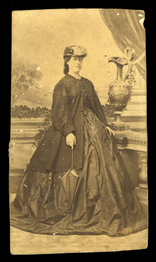 Woman with a large skirt and long jacket, holding an umbrella, and resting her hand beside a large urn