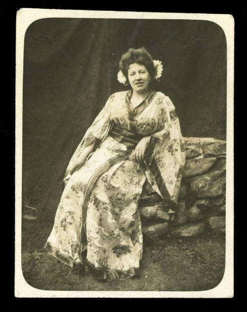 Woman wearing a kimono, with two flowers in her hair, sitting on a rock wall