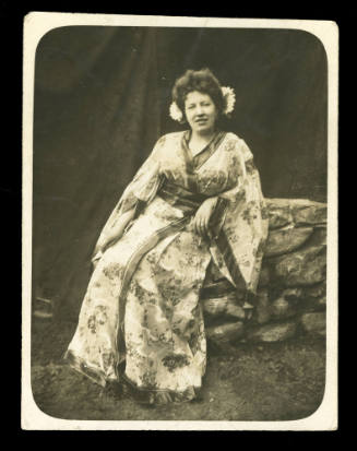 Woman wearing a kimono, with two flowers in her hair, sitting on a rock wall