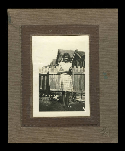 Woman with short hair, wearing a short white dress with cap sleeves, in front of a wooden fence with houses in the background