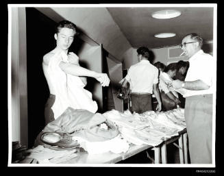 Man tries on shirt at table of clothing while man watches