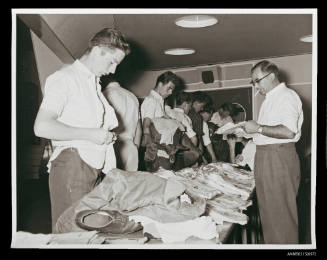 Young men examining clothes on tables and one trying some of them for a fit