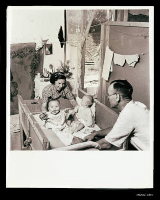 Woman and man look at two babies in large cot, displaced persons camp Europe