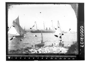 Possibly a Royal Mail steamer surrounded by yachts on Sydney Harbour with a small number of spectators seen seated on a rocky shore in the foreground