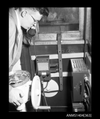 Photographic negative showing a man demonstrating a telephone installed on the bridge of a small vessel