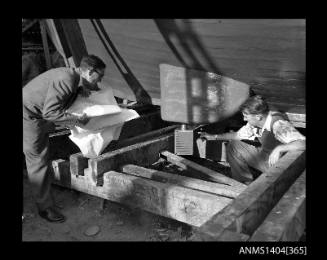 Photographic negative showing two men crouched underneath the hull of a vessel in dry dock at Kirribilli