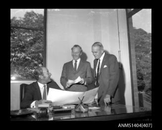 Photographic negative showing a group of three men looking at plans on an office desk