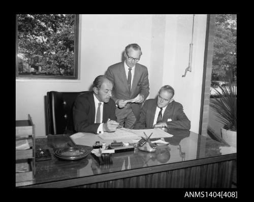 Photographic negative showing a group of three men looking at plans on an office desk